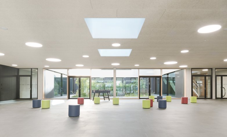 Großer, lichtdurchfluteter Innenraum mit modernen Designelementen. Die Decke verfügt über mehrere runde Leuchten und zwei große quadratische Oberlichter, die natürliches Licht hereinlassen. Vor einer Glasfassade mit Blick auf einen begrünten Außenbereich stehen farbenfrohe, zylinderförmige Sitzhocker in verschiedenen Farben wie Blau, Rot und Grün. Der Raum wirkt offen und minimalistisch, ideal für Begegnungen, Veranstaltungen oder gemeinschaftliche Aktivitäten.